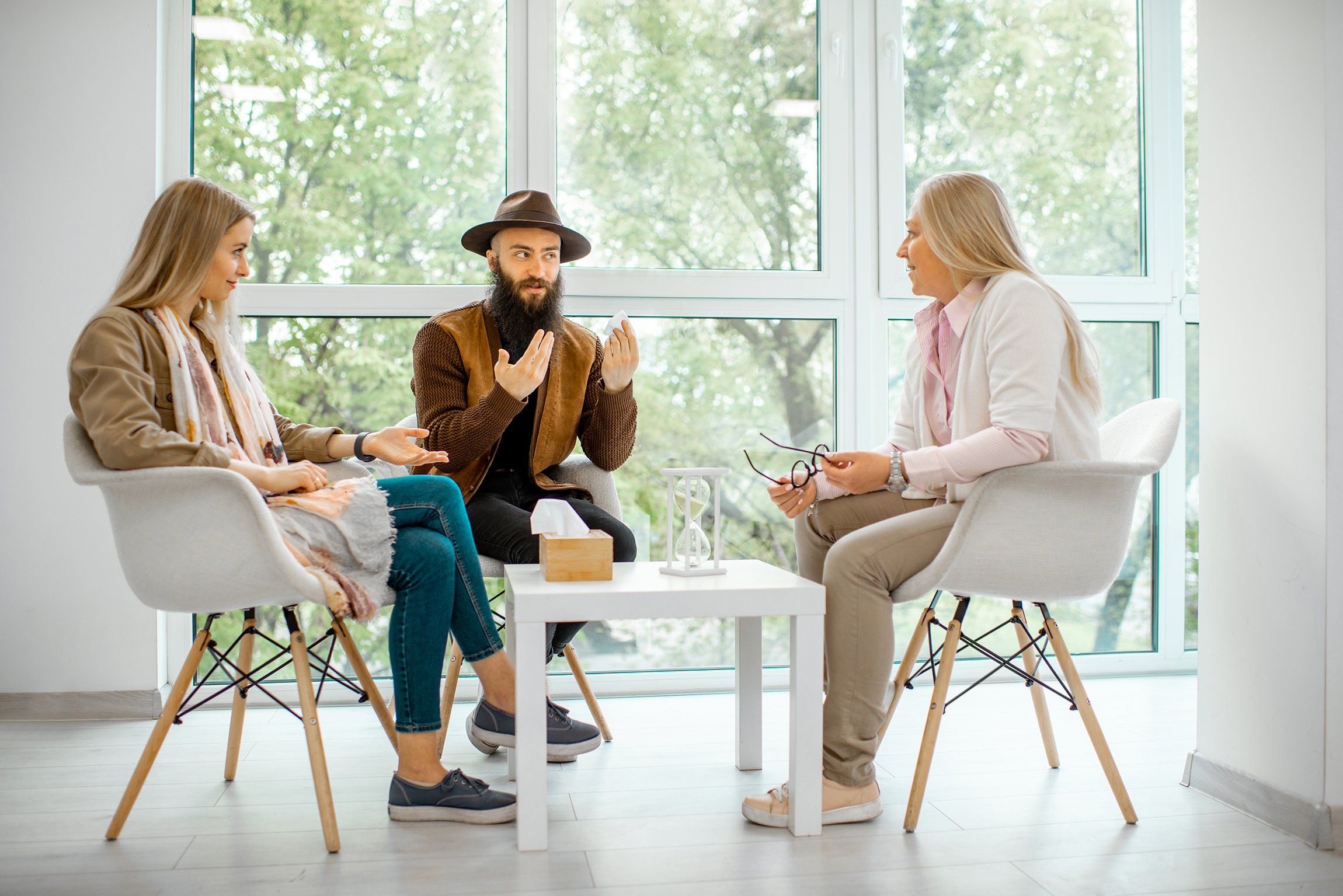 Three adults sit in a bright room having a casual discussion around a small white table, the bearded man speaking while two women listen.