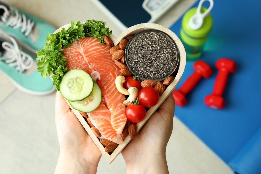 Hand holding a heart-shaped wooden bowl filled with salmon, cucumber, greens, nuts, tomatoes and chia seeds for a healthy meal.