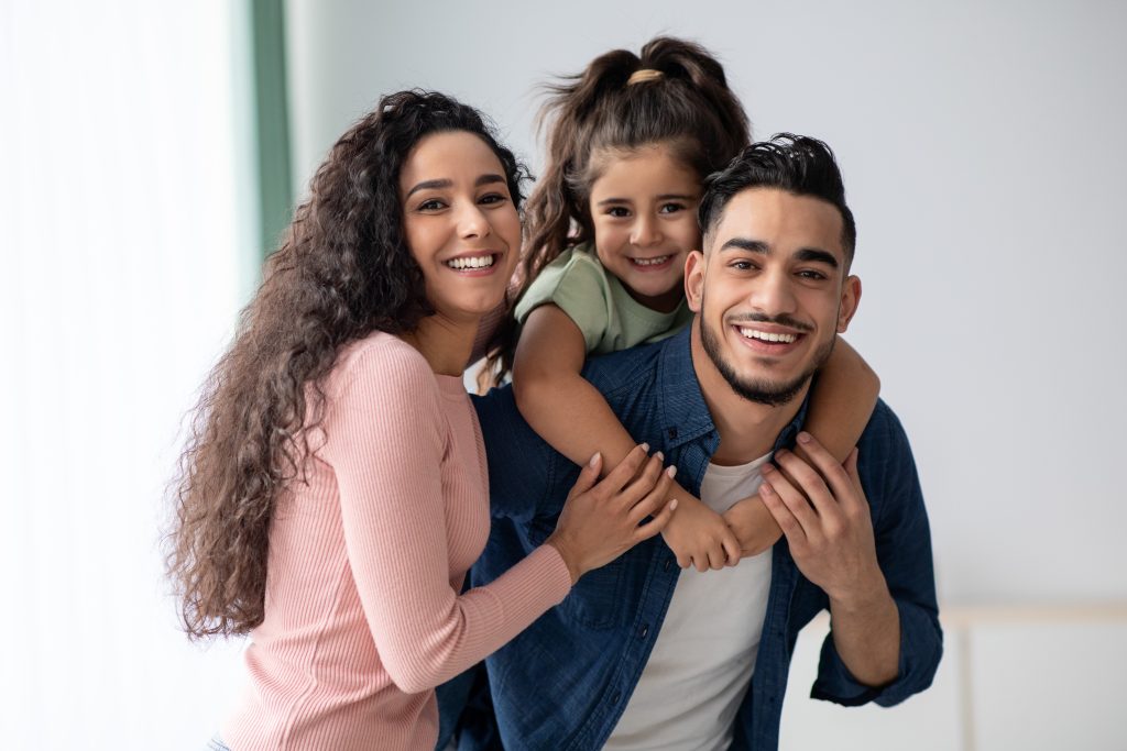 Smiling family of three posing indoors: a woman with long curly hair on the left, a young girl on a man's shoulders, all looking at the camera and smiling.