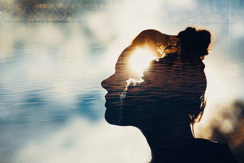 Double exposure of a woman's silhouette over a lake with sunlight behind, creating a dreamy, reflective mood.