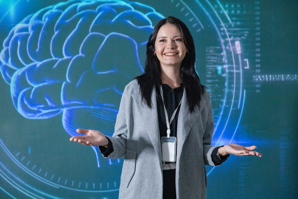 Smiling woman in a gray blazer presents with open hands in front of a blue neon brain backdrop.